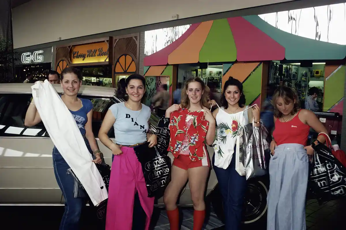 Five young women in 1980s fashion stand proudly in front of a shopping mall storefront, each holding shopping bags or new…