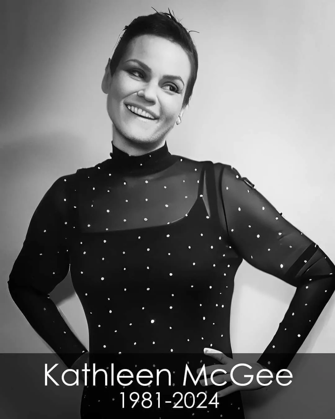 A black and white portrait of Kathleen McGee smiling with her hands on her hips, wearing a polka-dotted, sheer-sleeved top…