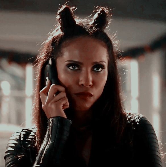 a woman with dark hair styled into two small horns holds a phone to her ear, wearing a black leather jacket, with a serious…