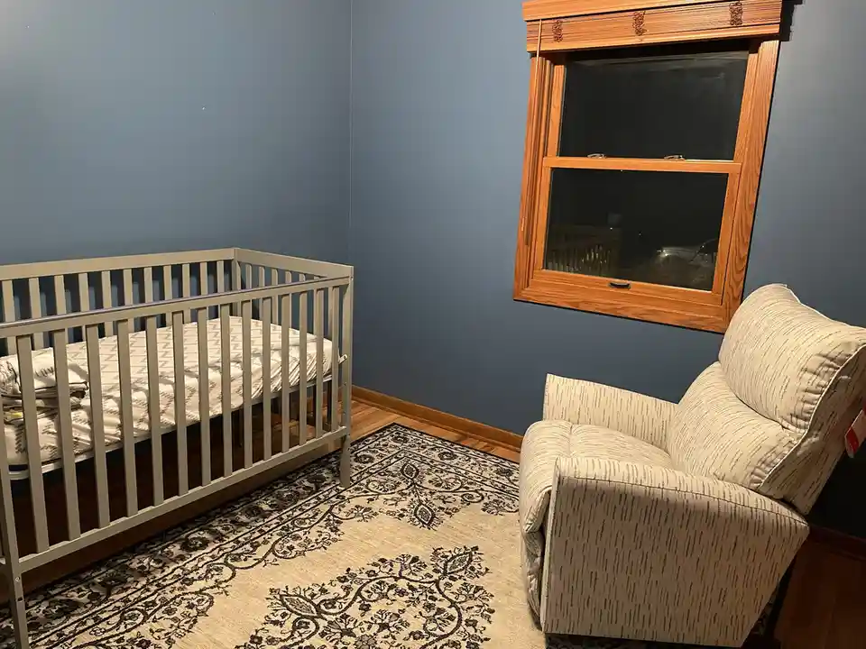 A cozy nursery with a gray crib, a patterned rug, and a recliner positioned near a wooden window. The room has blue walls…