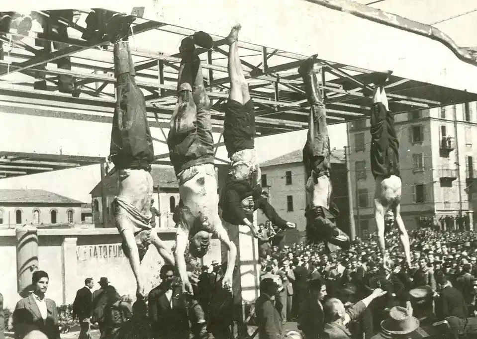 A black-and-white historical photograph shows five men hanging upside down from a metal frame in front of a large crowd. The…