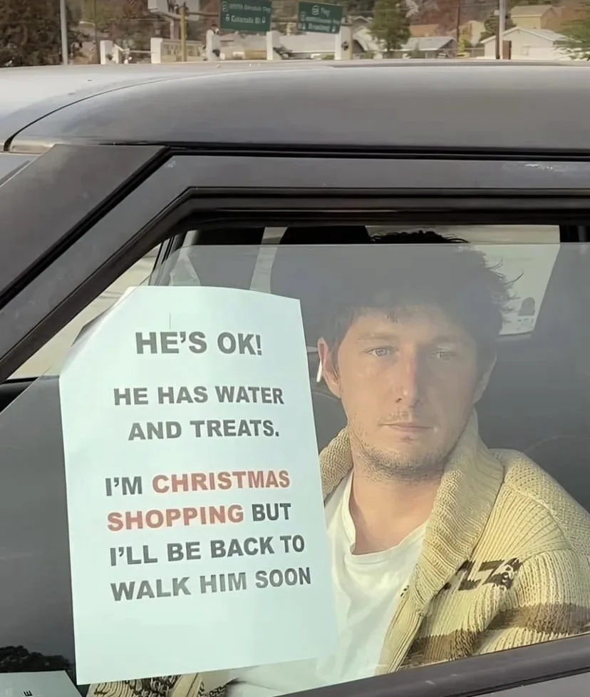 A man sits in the driver's seat of a car, looking out the window, with a sign taped to the passenger side window. The sign…