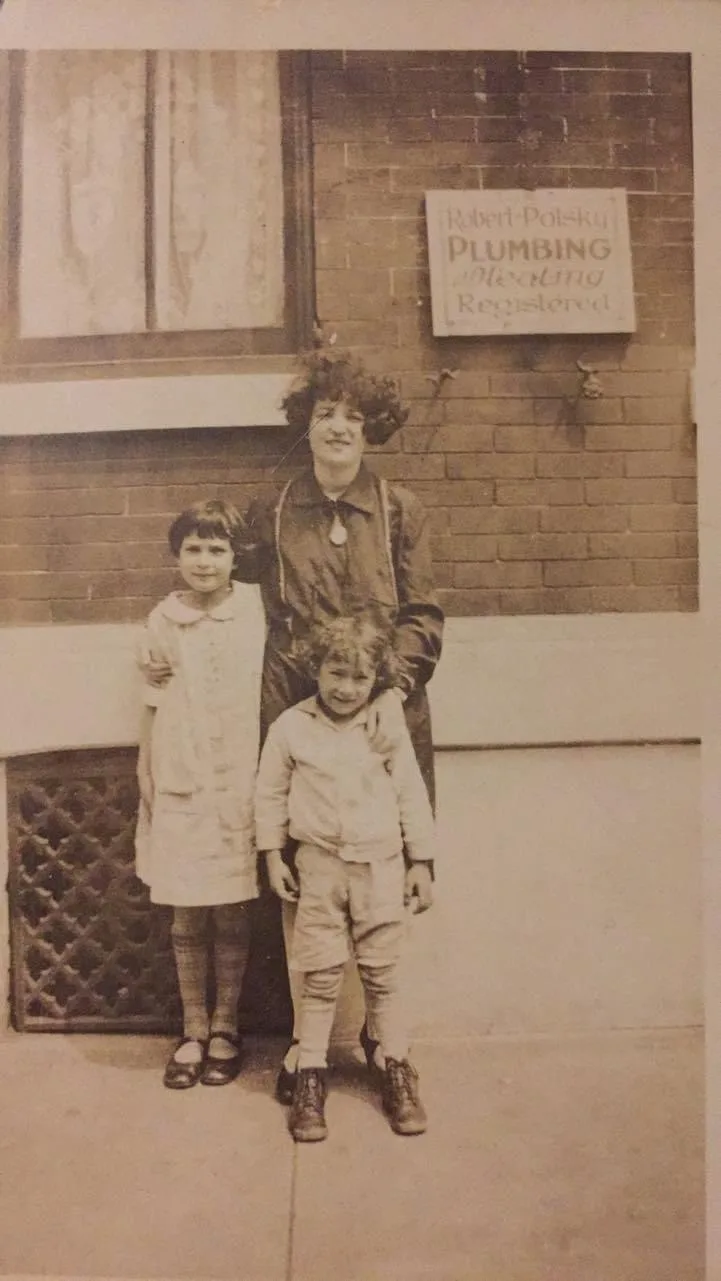 A sepia-toned vintage photo shows a woman with curly hair standing between two children in front of a brick wall. A sign…