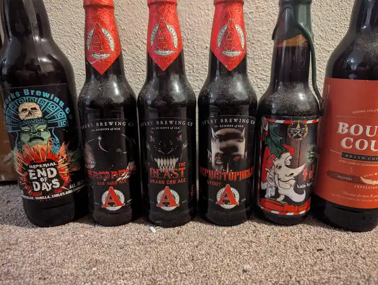 Six dark beer bottles are lined up on a carpeted floor, with five from Avery Brewing Co. featuring demonic-themed labels…