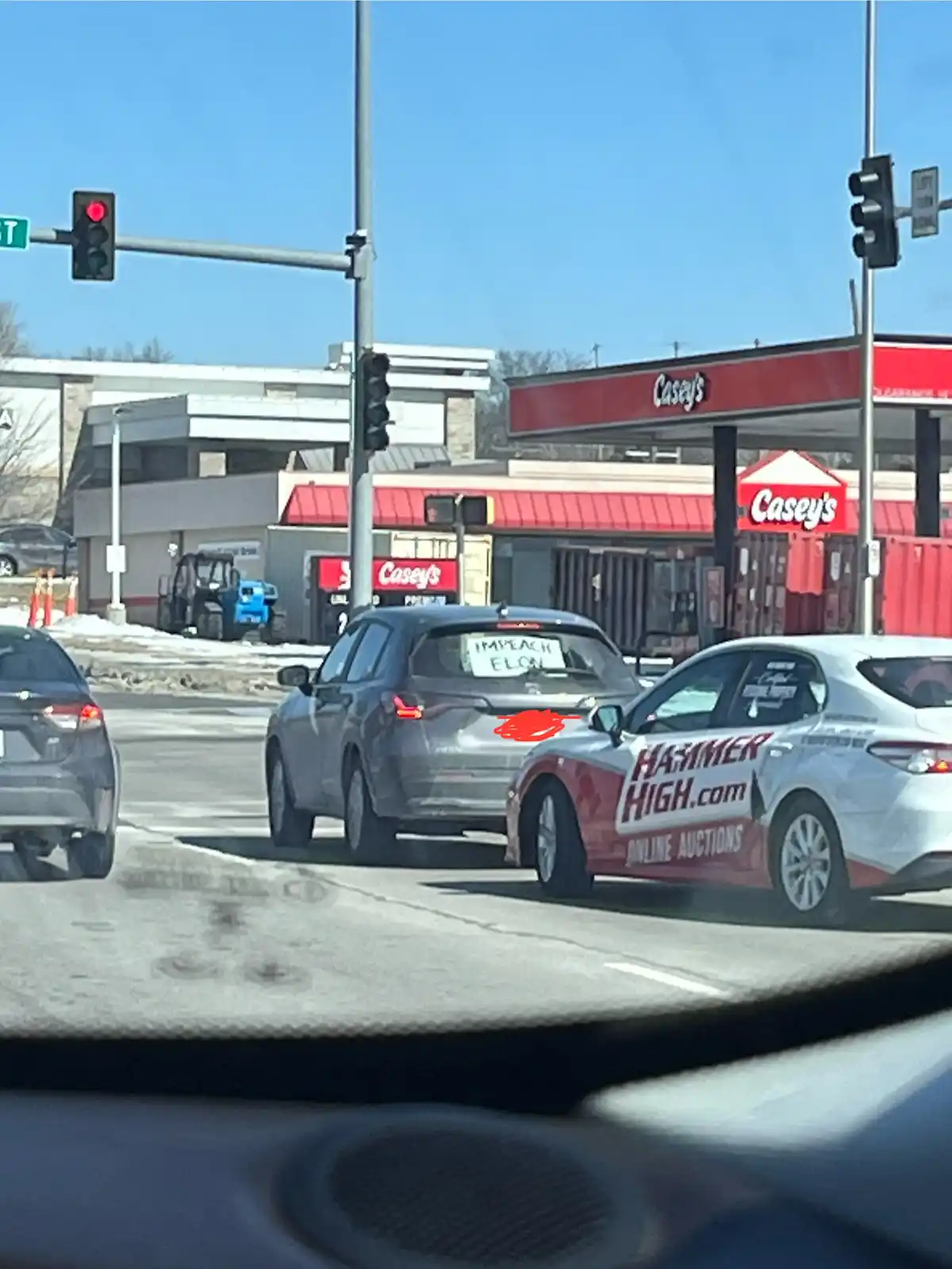 A gray SUV is stopped at a red traffic light with a handwritten sign in its rear window that reads 'IMPEACH ELON'. The car…