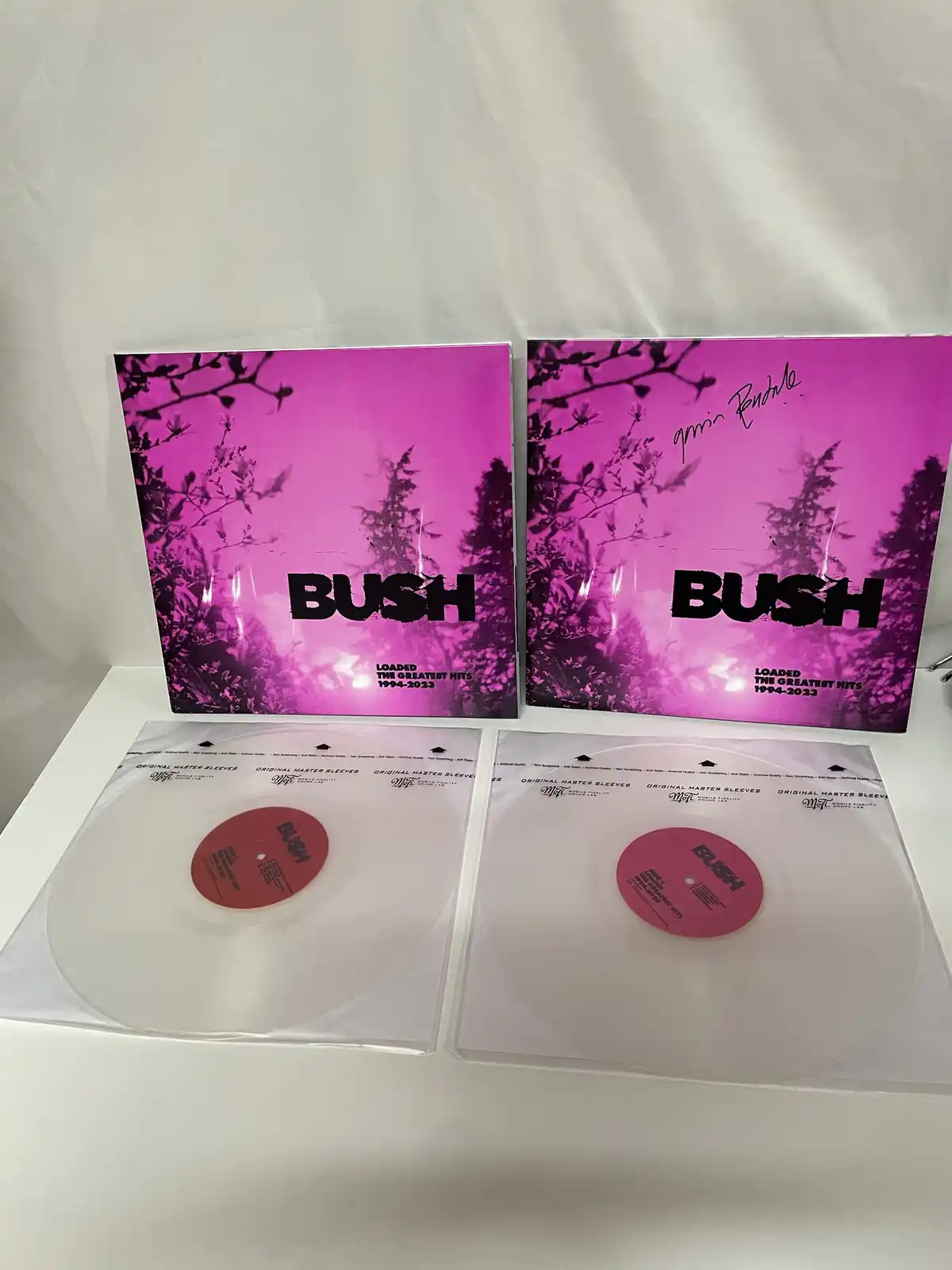 Two identical pink album covers for 'BUSH: LOADED THE GREATEST HITS 1994-2023' stand behind two clear vinyl records, one…