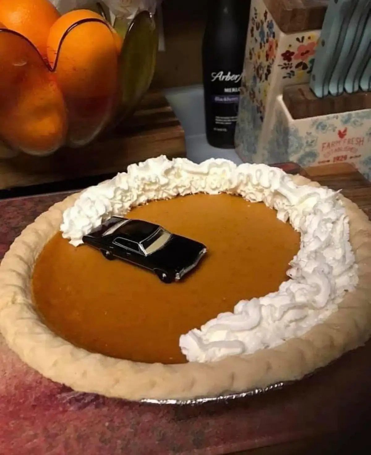 A close-up photo shows a pumpkin pie with whipped cream around the edge, and a small black toy car placed in the center of…