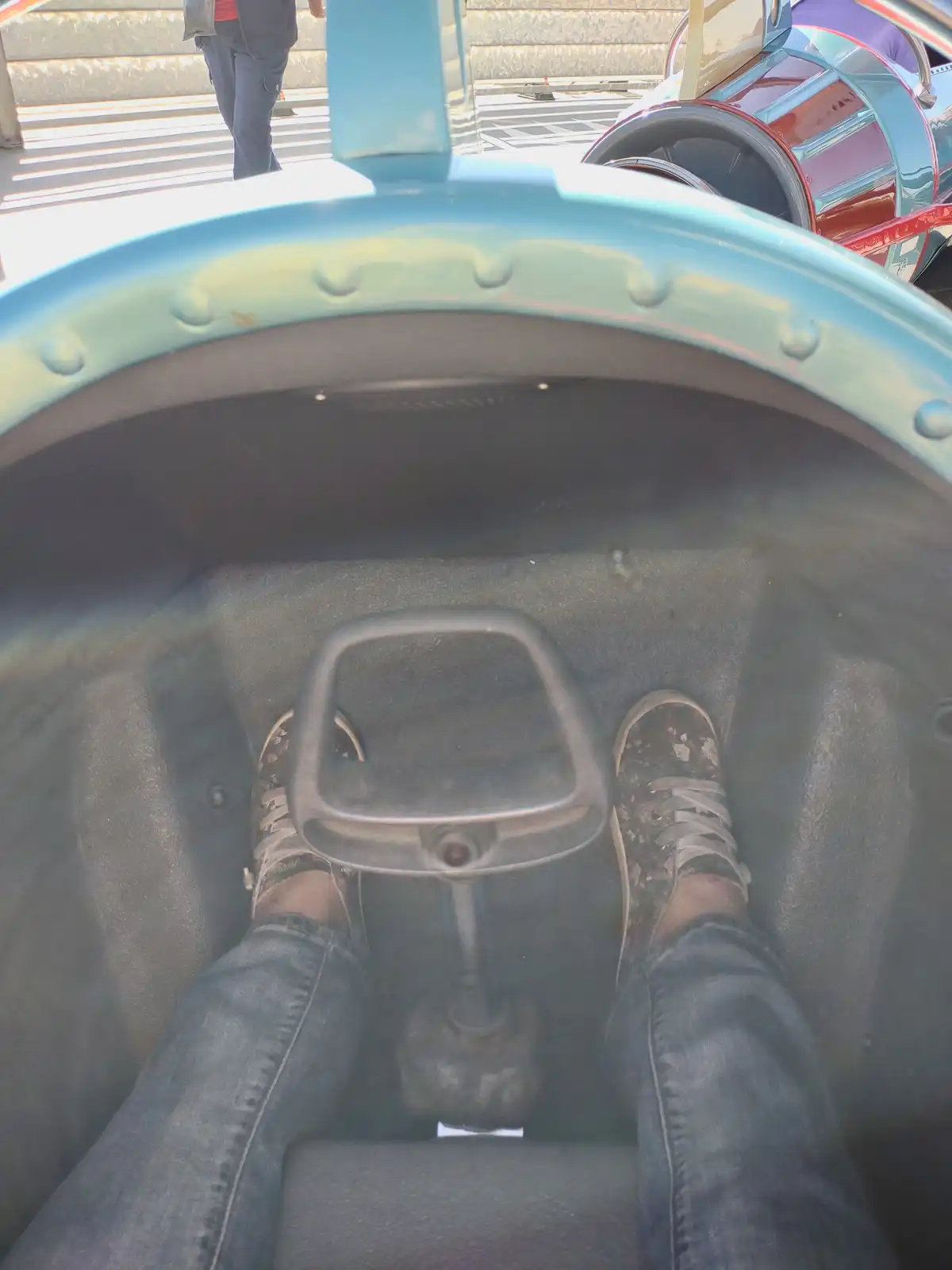 A first-person view from inside a small, colorful ride vehicle, showing a person's feet in sneakers and jeans positioned on…