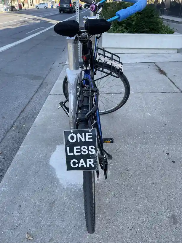 A blue bicycle is parked on a sidewalk next to a street, viewed from behind. Attached to the rear of the bike is a black…