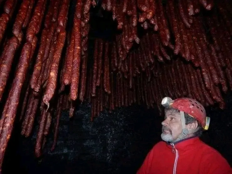a man wearing a red helmet with a headlamp and a red jacket stands in a dark cave, looking up at hundreds of large…