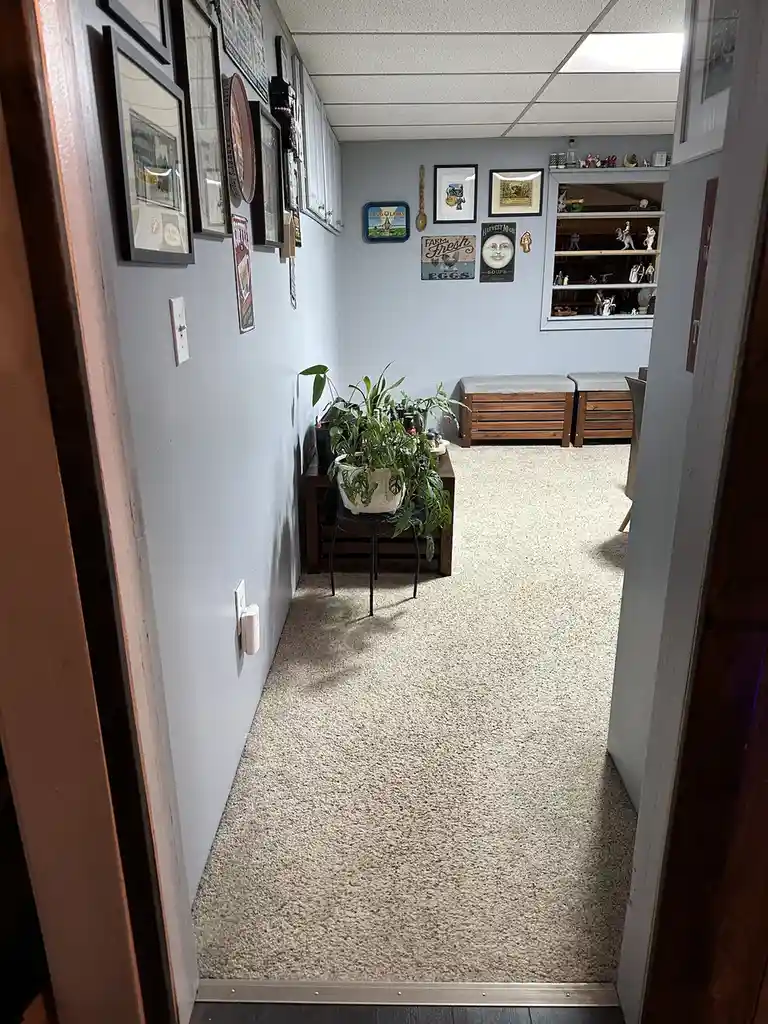 A view from a doorway into a narrow hallway with light blue walls, carpeted floor, and numerous framed pictures and…