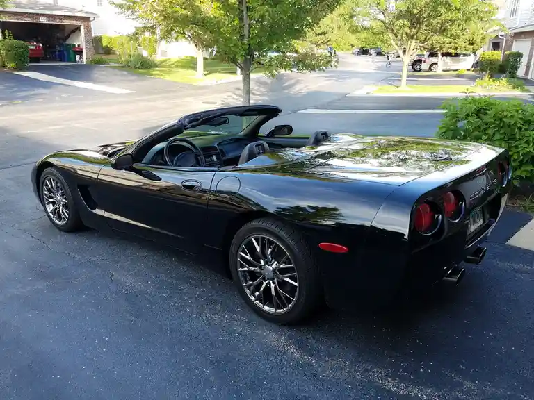 A black Chevrolet Corvette convertible is parked on a residential street, its top down, reflecting trees and sky. The car…