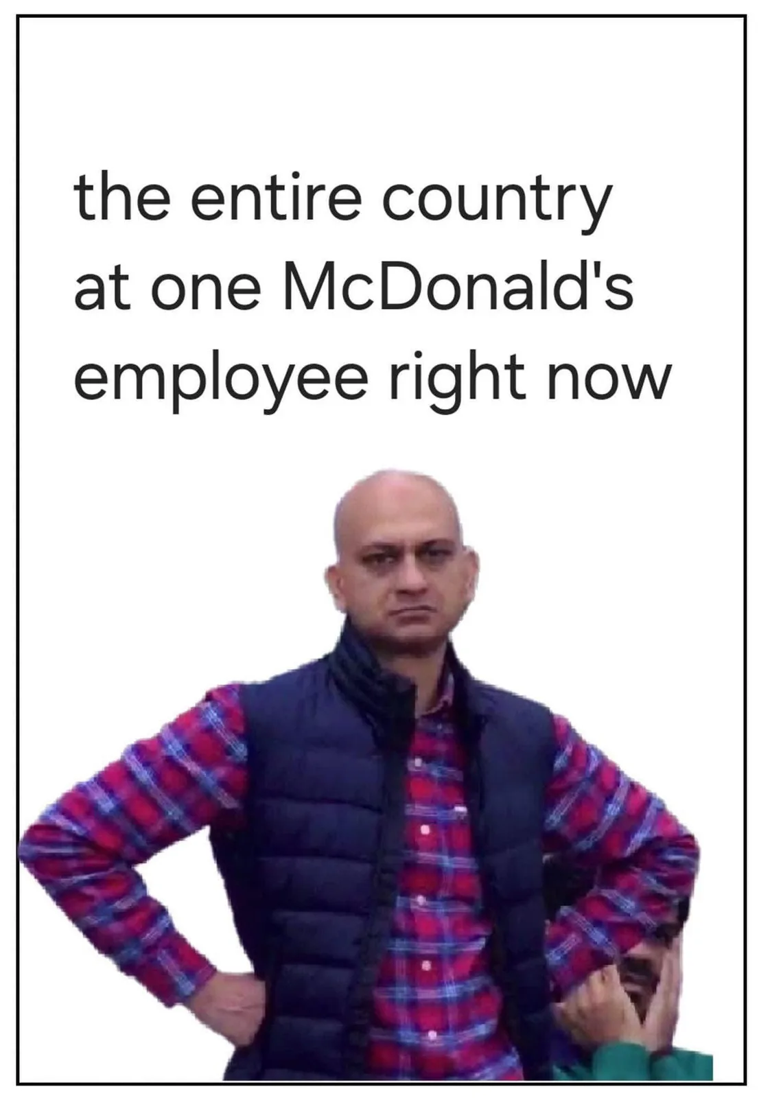 A bald man in a red plaid shirt and black vest stands with hands on hips, looking sternly at the viewer. Behind him, another…