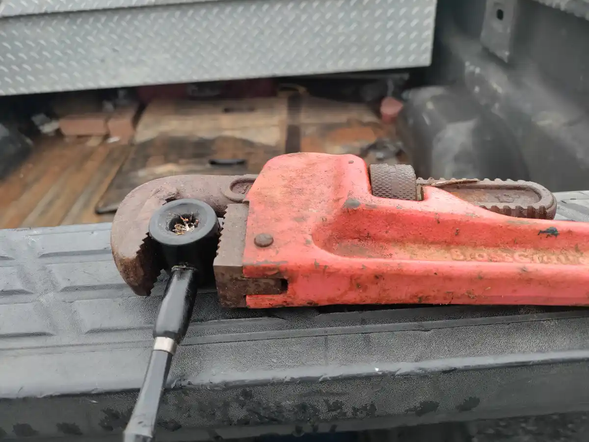 A red, rusty pipe wrench rests on a black textured surface, with a black pipe smoking from its jaws. The overlaid text reads…