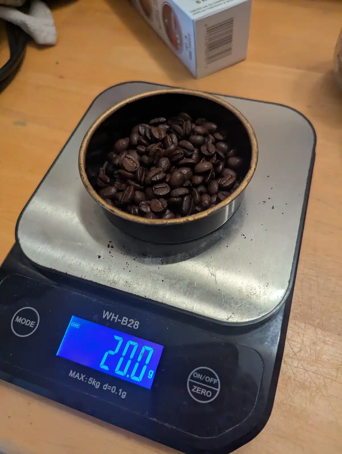 A digital kitchen scale displays '20.0 g' while holding a small metal cup filled with roasted coffee beans. The scale is on…