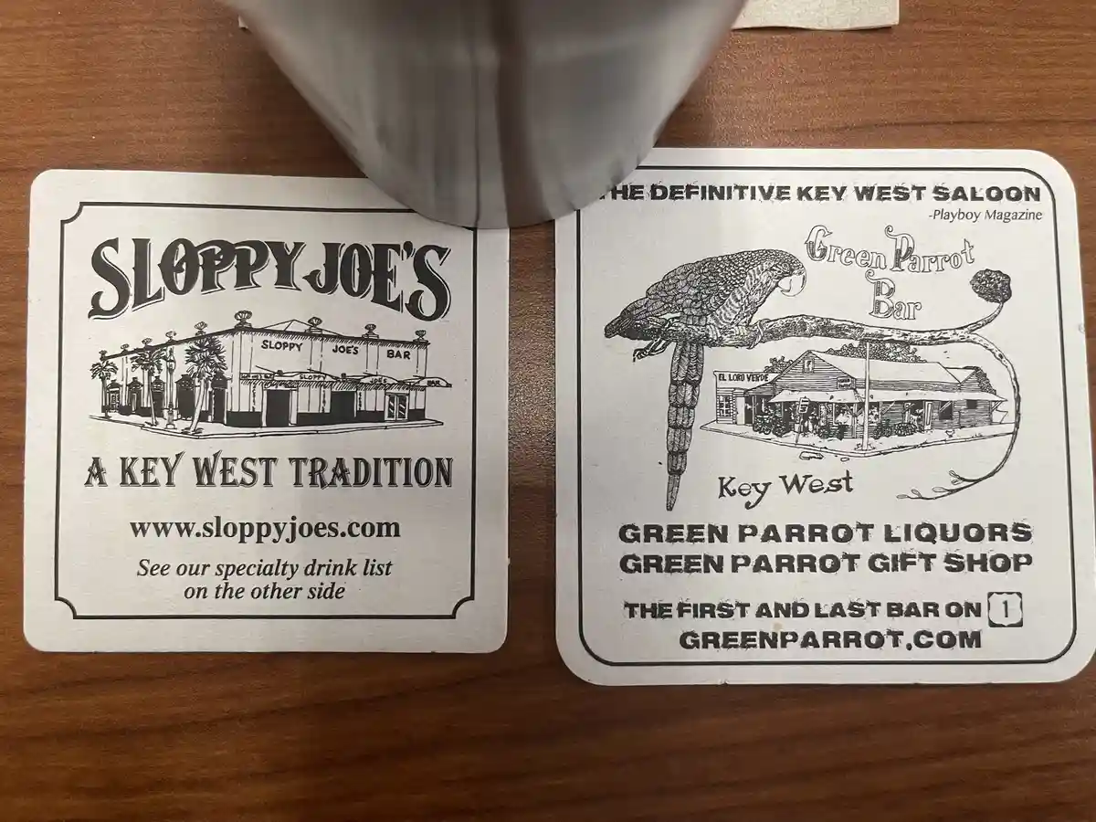 Two coasters for Key West bars, Sloppy Joe's and Green Parrot Bar, sit side by side on a wooden table with a gray mug…
