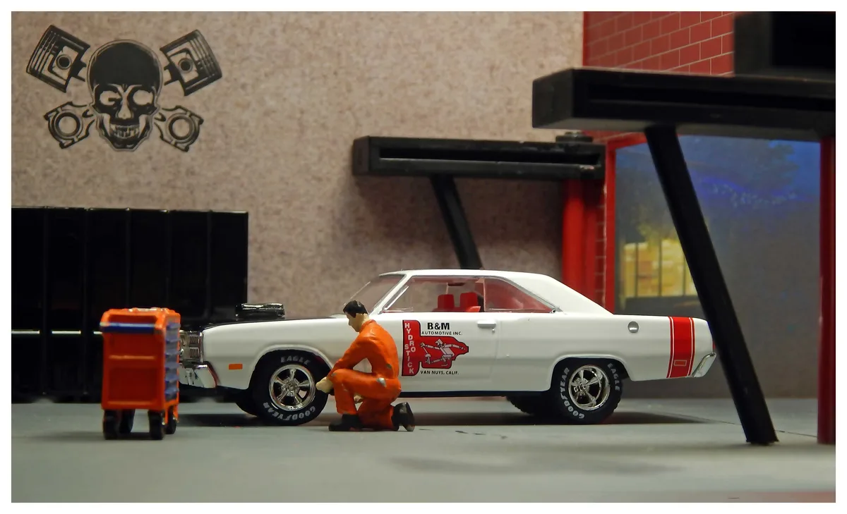 A miniature diorama shows a small figurine mechanic in an orange jumpsuit kneeling beside a white classic muscle car with…
