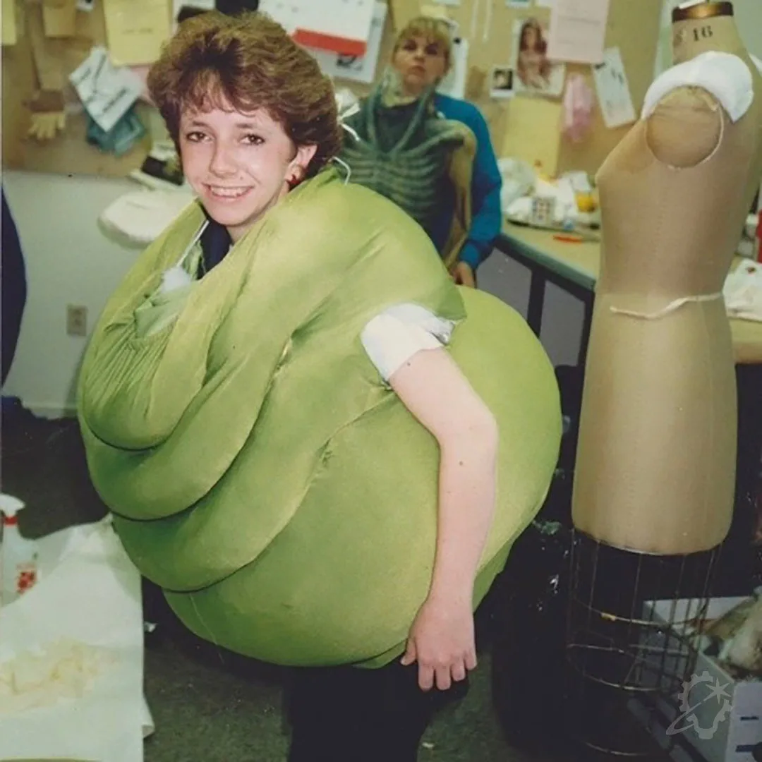 A young woman in a costume workshop is wrapped in a large, green, bulbous costume that obscures her body, leaving only her…