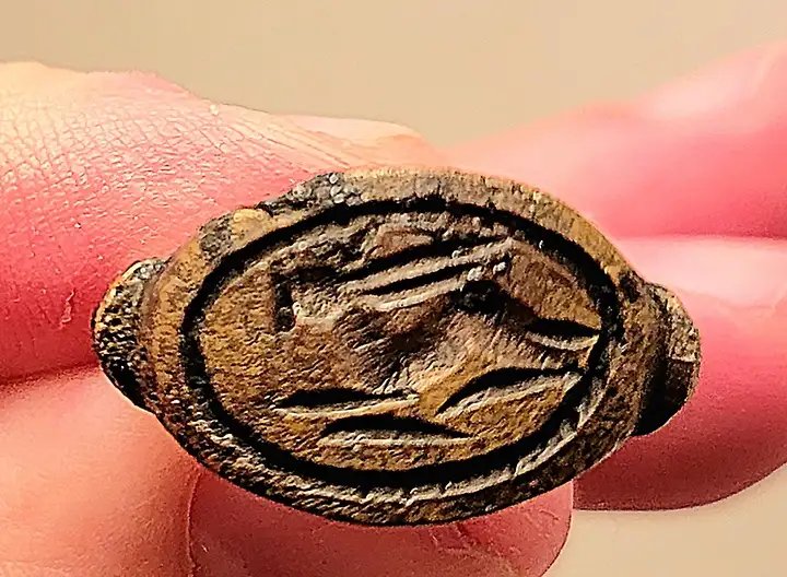 A close-up photo shows a small, oval, bronze-colored ring held between fingers, featuring an embossed, stylized face with…