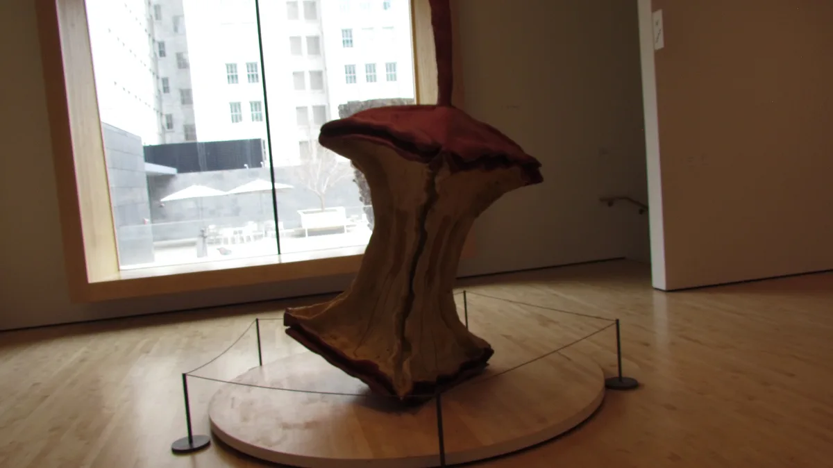 A large, abstract sculpture resembling a sliced apple core with red fabric on top sits on a circular wooden platform in a…