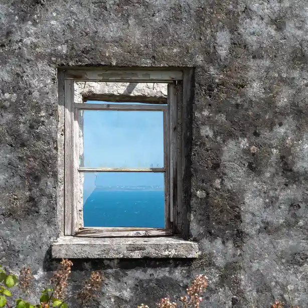 A weathered, broken wooden window set in a rough, gray stone wall frames a view of a bright blue ocean and sky. The glass is…