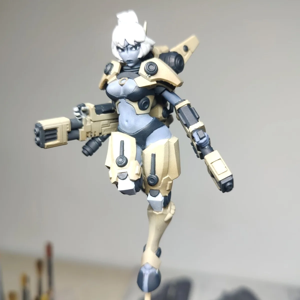 A painted action figure of a female mech pilot with white hair, beige and black armor, and a weapon in her right hand, posed…