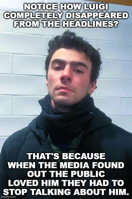 A man with dark hair and a black hooded jacket stands against a white tiled wall, looking directly at the camera with a…