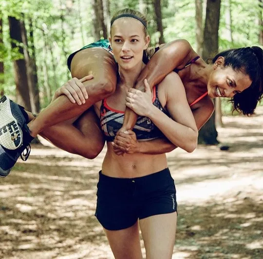Two fit women in workout clothes are in a forest; one carries the other on her back while smiling. The carried woman has a…