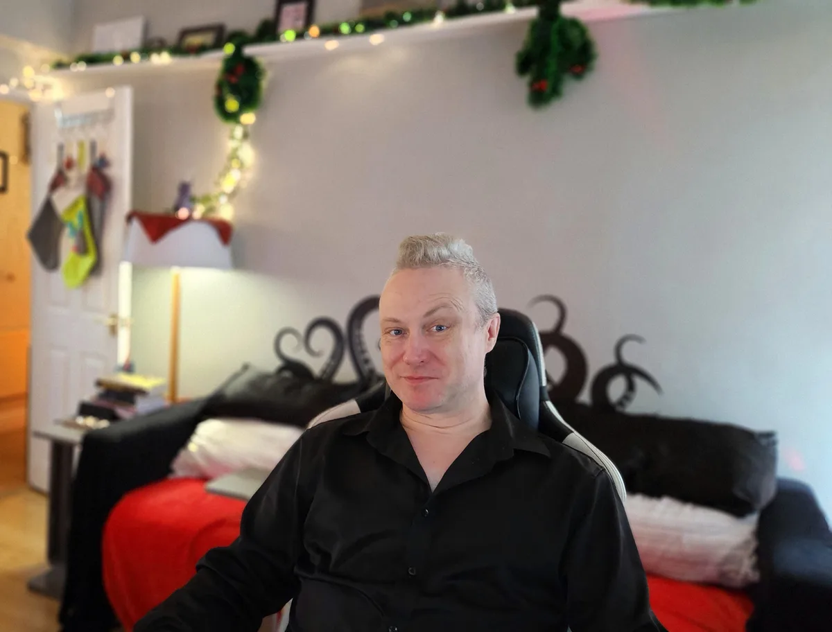 A man with gray hair sits in a black gaming chair in a room decorated with Christmas lights and garlands. Behind him, a…