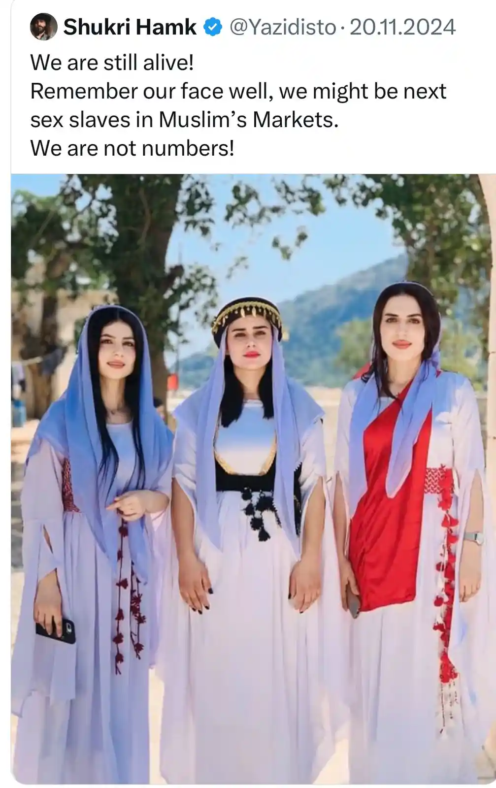 Three women in traditional Yazidi attire stand together outdoors under trees. The overlaid text reads: 'We are still alive!…