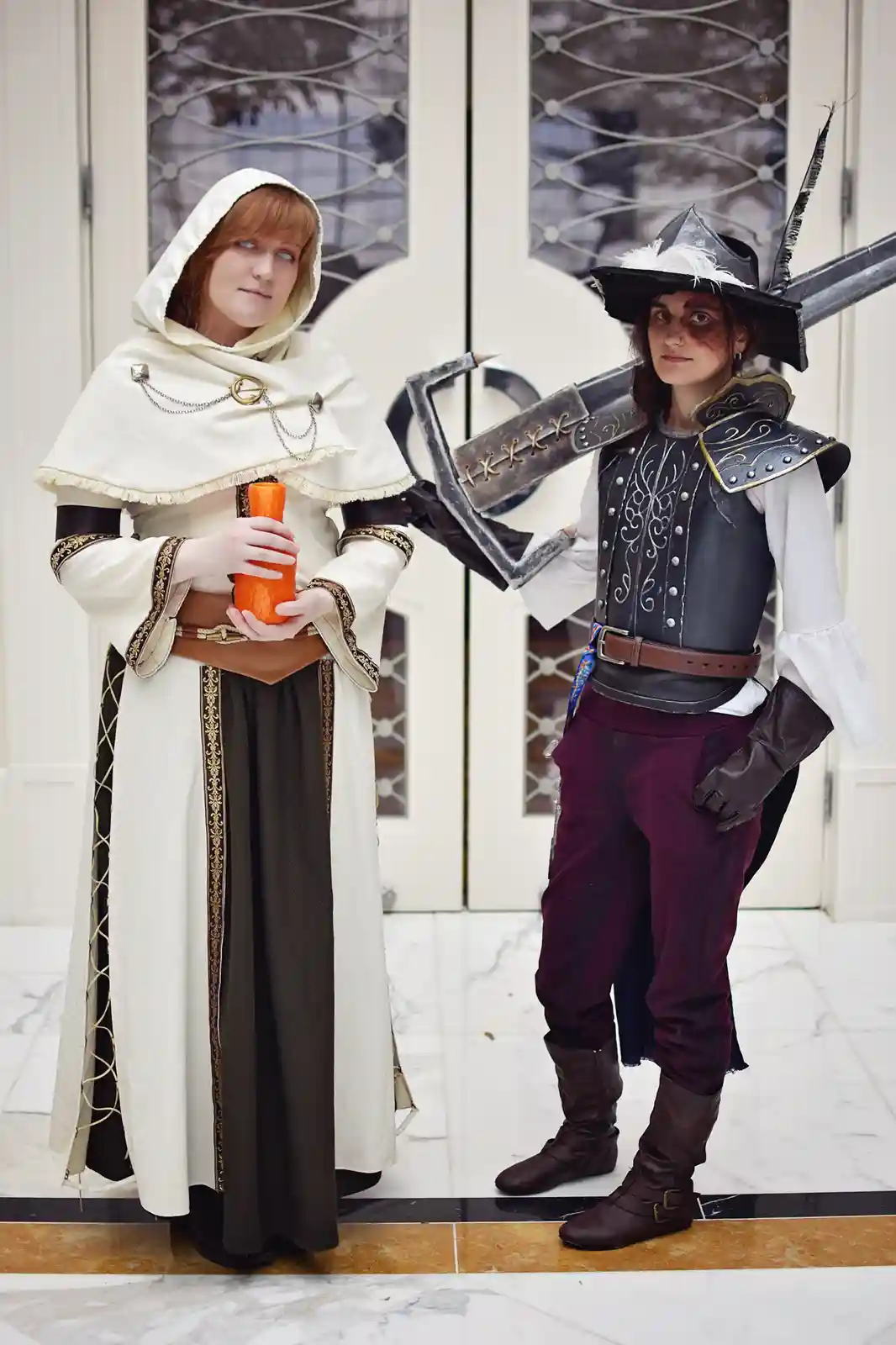 Two people in elaborate fantasy costumes stand side by side. The person on the left wears a white hooded robe with gold trim…