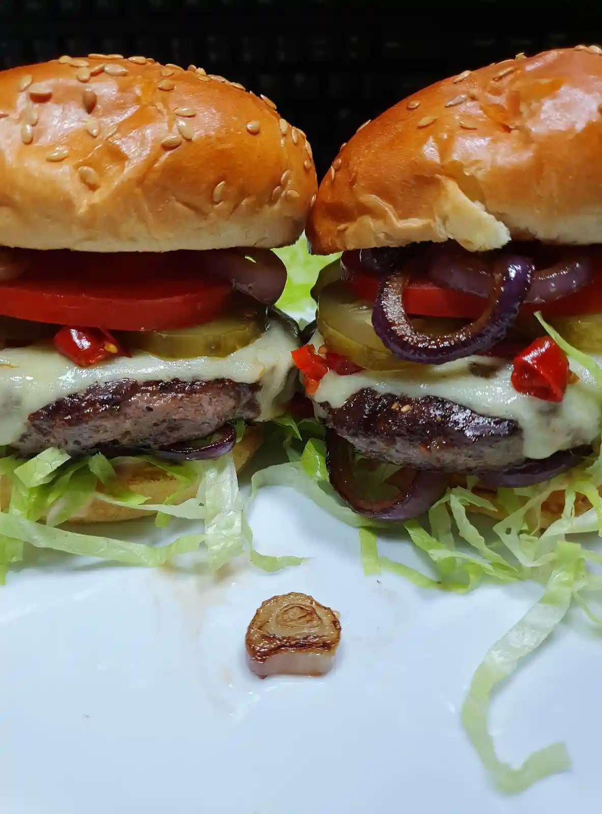 Two gourmet-looking cheeseburgers with sesame seed buns, lettuce, tomato, pickles, onions, and melted cheese sit side by…