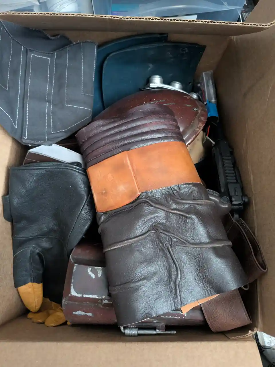 A cardboard box filled with various worn cosplay props, including a large brown leather glove with an orange band, a black…