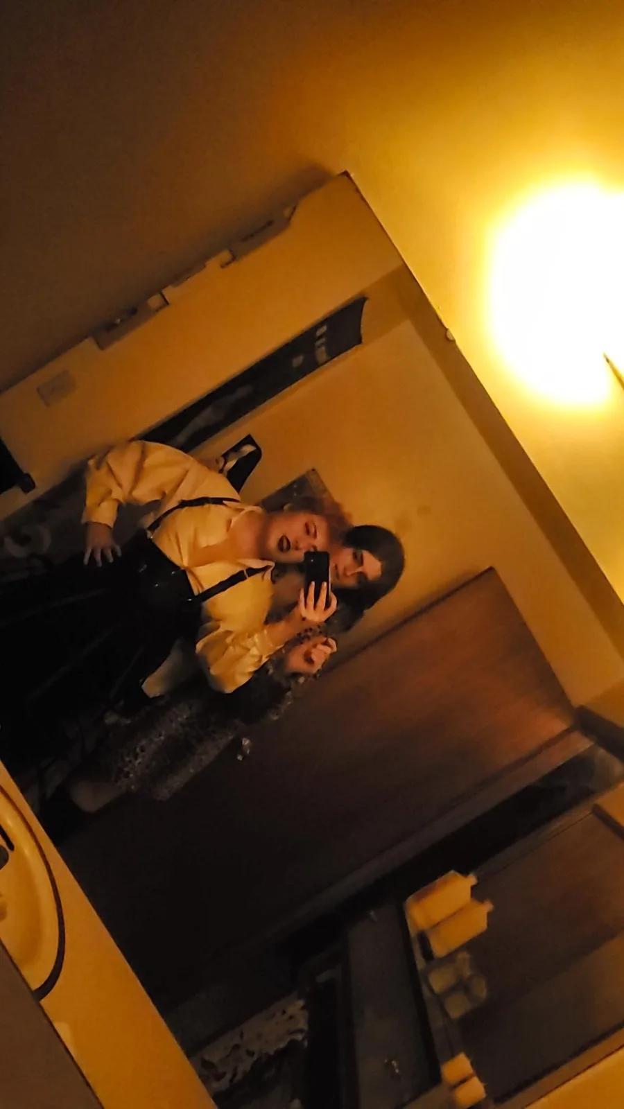 A person with dark hair and a yellow shirt is taking a selfie in a mirror in a dimly lit bathroom, holding a phone. The…