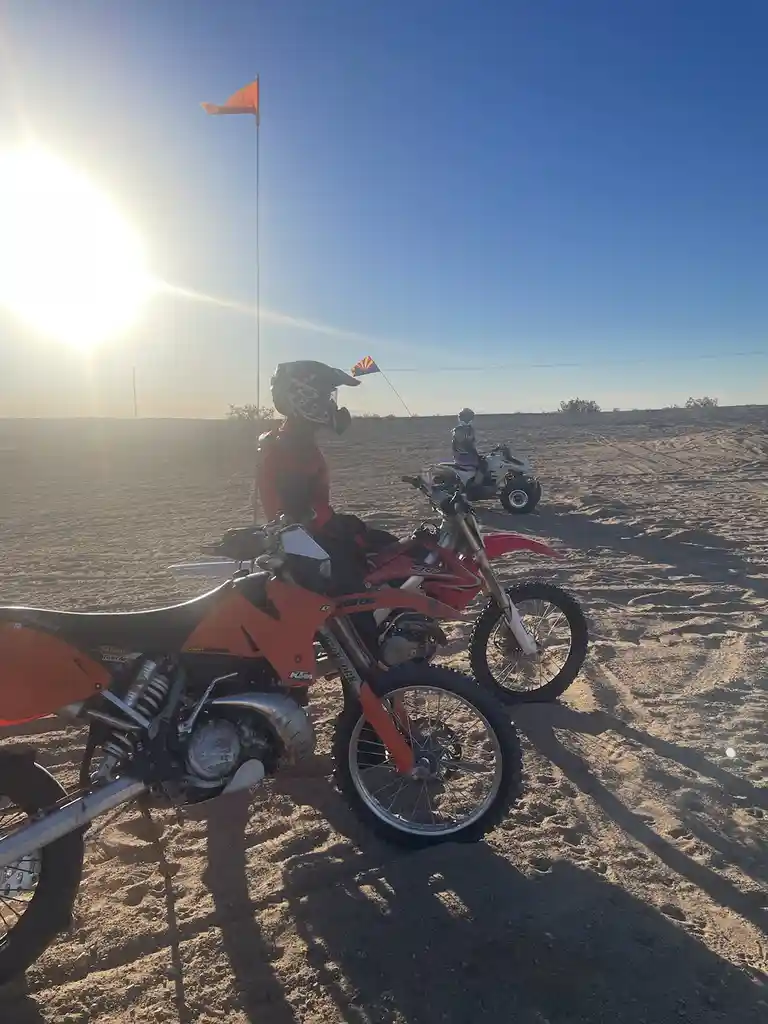 Two people in full motocross gear sit on dirt bikes in a vast, sunlit desert. One rider is in the foreground on an orange…