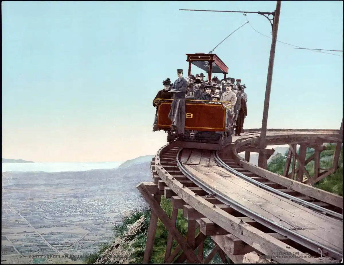 An old, colorized photograph shows a crowded, open-air cable car labeled '9' on a wooden trestle track high above a valley…