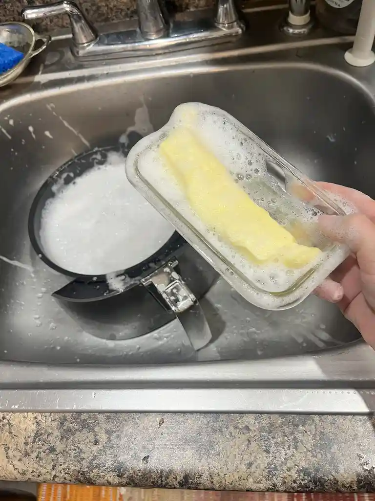 A hand holds a clear glass dish filled with soapy water and a yellow, butter-like soap bar, lifted above a stainless steel…