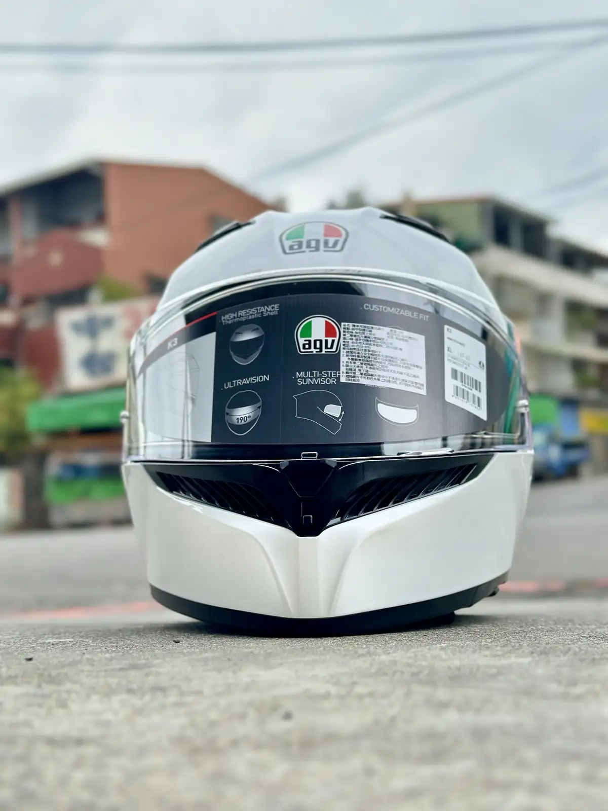 A white AGV motorcycle helmet sits on pavement, its visor still covered by a black sticker detailing features like 'HIGH…