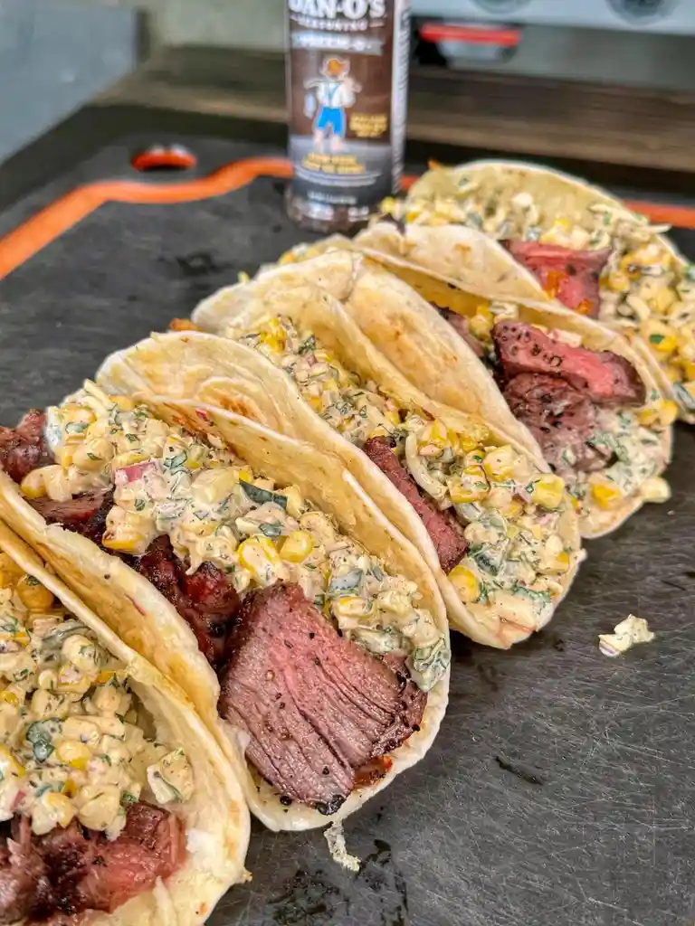 A row of four soft tacos filled with sliced medium-rare steak and creamy corn salsa sit on a black cutting board. In the…