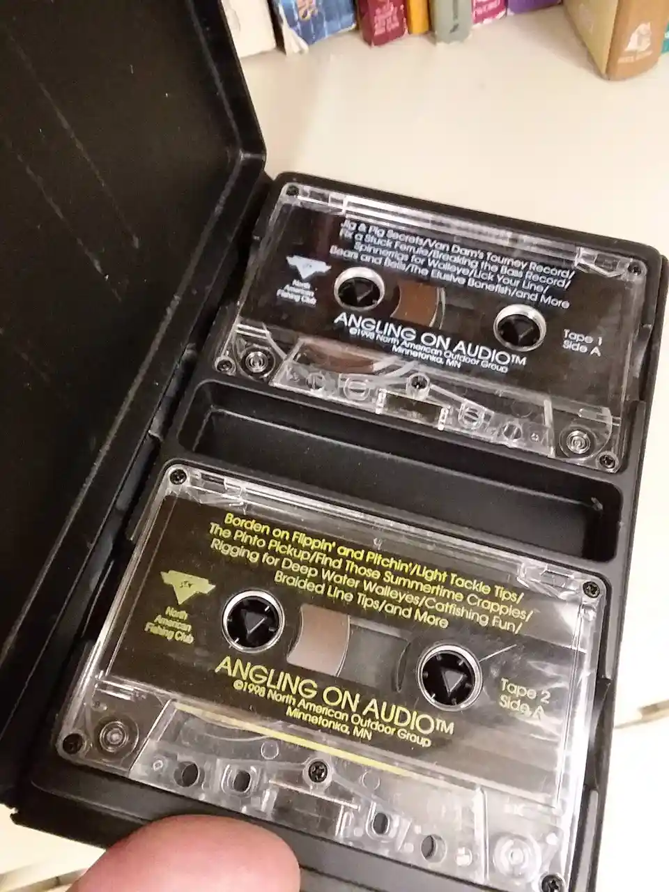 Two vintage audio cassette tapes labeled 'Angling on Audio™' from 1998 are shown in their open black case. The top tape…