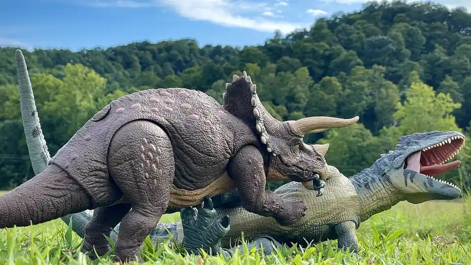 Two plastic dinosaur toys, a grey triceratops and a blue-green theropod, are staged in a grassy field with a forested hill…