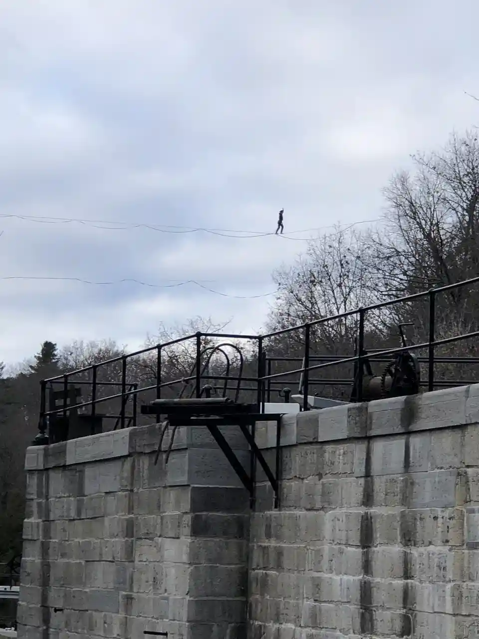 A person is walking on a tightrope strung high above a stone canal lock structure, with bare trees and a cloudy sky in the…