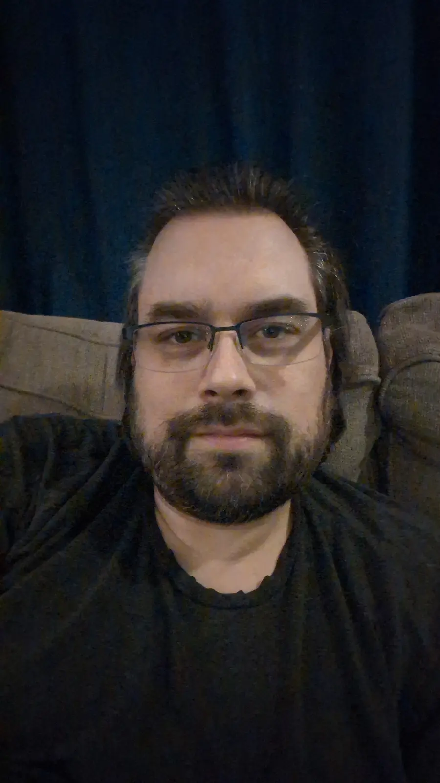 A man with glasses and a beard sits on a couch against a dark curtain, looking directly at the camera with a neutral…