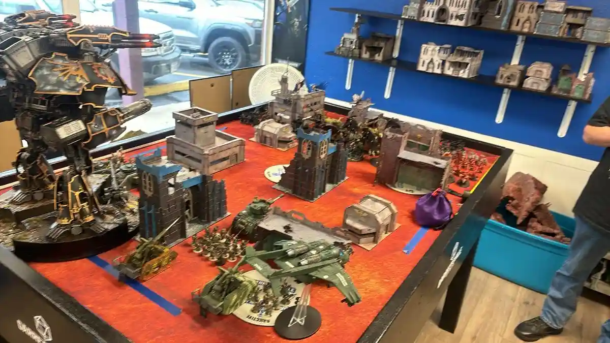 A large, detailed tabletop wargaming setup featuring miniature war machines, buildings, and armies on a red carpeted table…