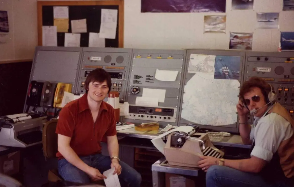 Two men sit at a large, retro control panel filled with dials, switches, and maps. The man on the left, in a red shirt…