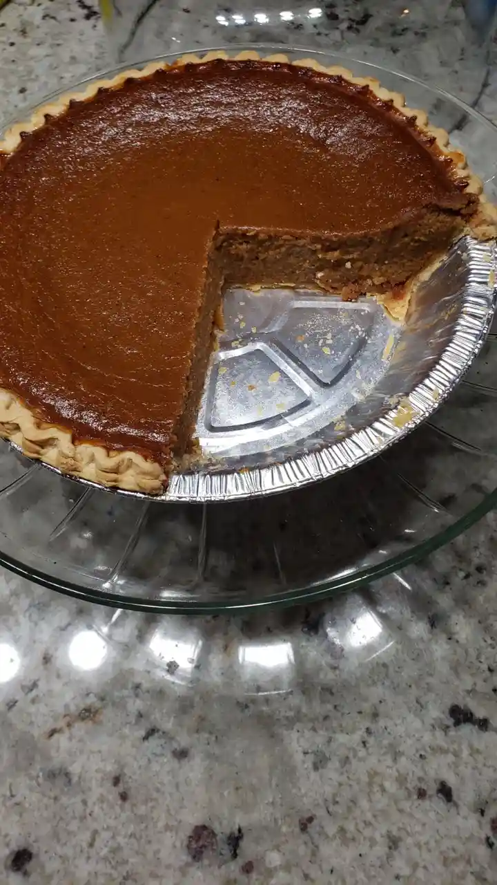 A close-up photo shows a pumpkin pie in a foil pie tin, sitting on a glass plate atop a granite countertop. A single slice…