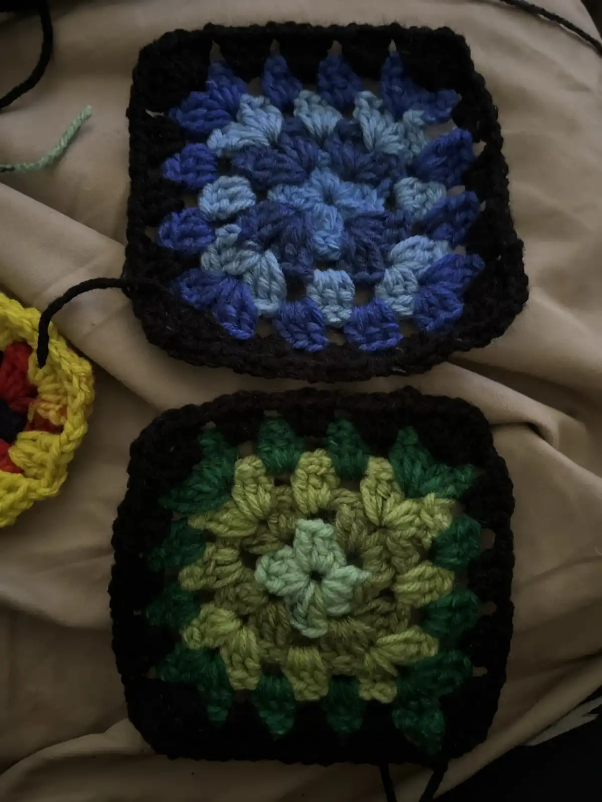 Two handmade crocheted granny squares are shown, one with blue and black yarn resembling a flower, the other with green and…