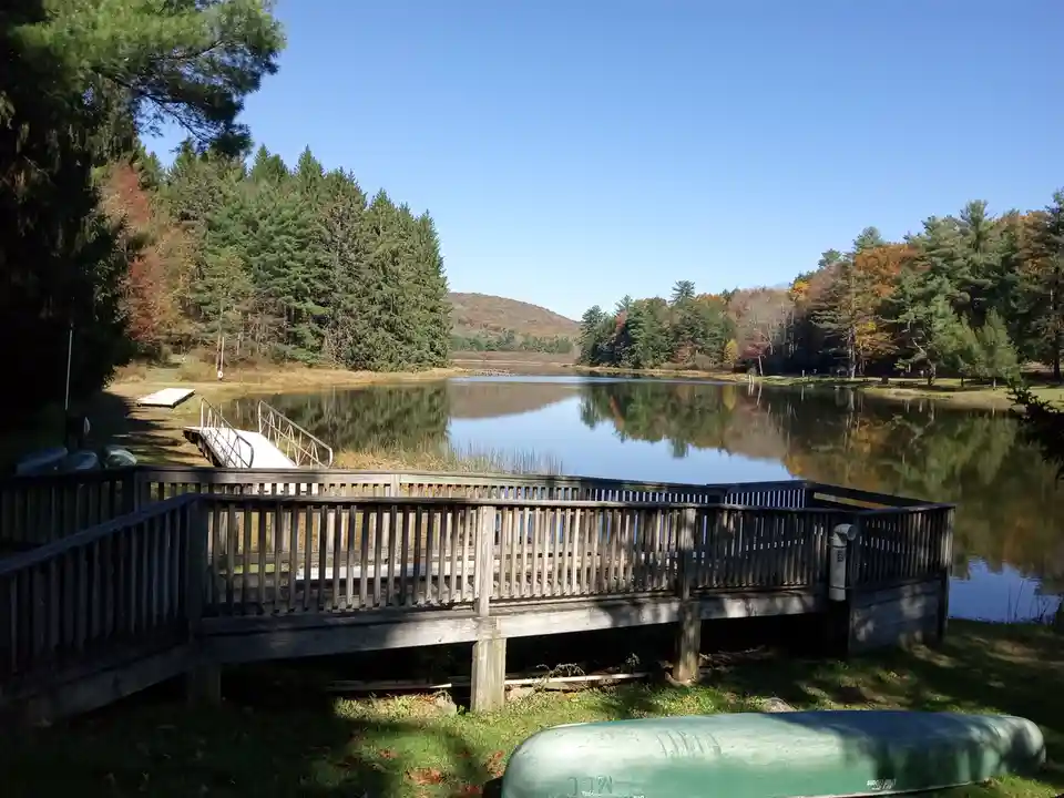 A serene lake surrounded by autumn-colored trees under a clear blue sky, with a wooden dock and a green canoe in the…