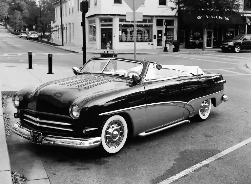 A black-and-white photo shows a vintage 1951 Hudson Hornet convertible parked on a street corner. The car has a glossy black…