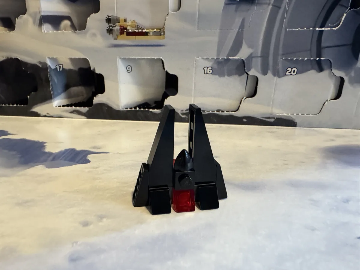 A small black and red Lego X-wing fighter model stands on a white surface in front of a grey cardboard box with numbered…